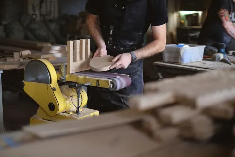 Unrecognizable male artisan using belt and disc sander to shape wooden detail while working near anonymous coworker in workshop with stack of wooden boards